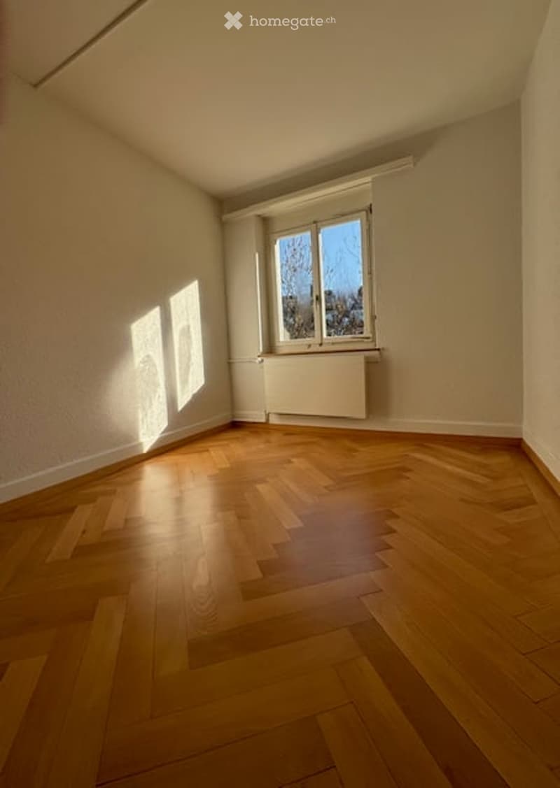 Empty room with wooden floor, white walls, large window, and radiator below the window