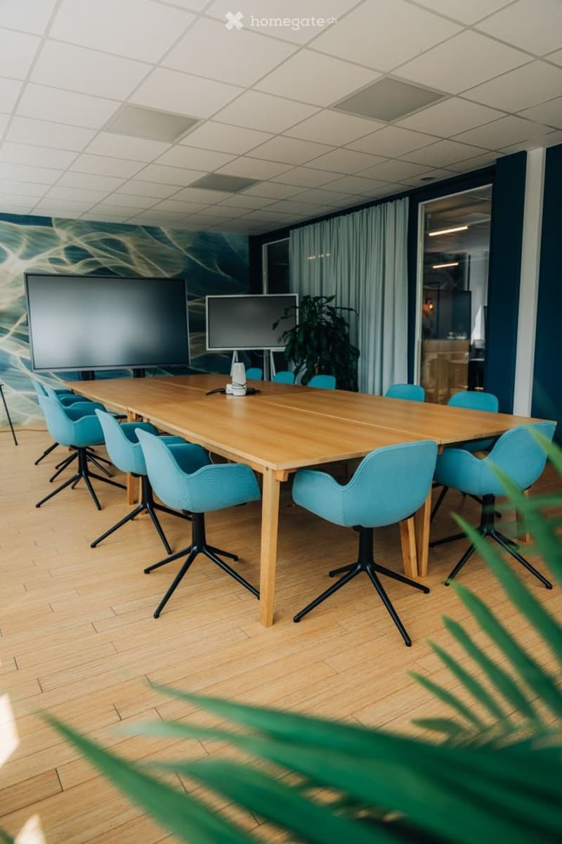 Furnished Room. Modern conference room with a long wooden table surrounded by blue chairs, two large screens mounted on the wall, and a potted plant near the window.
