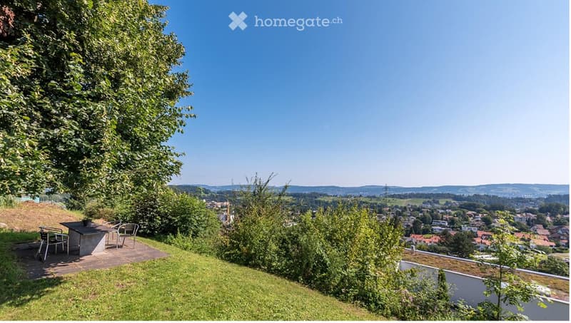 A serene garden or outdoor area with a table and chairs, surrounded by lush greenery and offering a panoramic view of a town and distant hills.