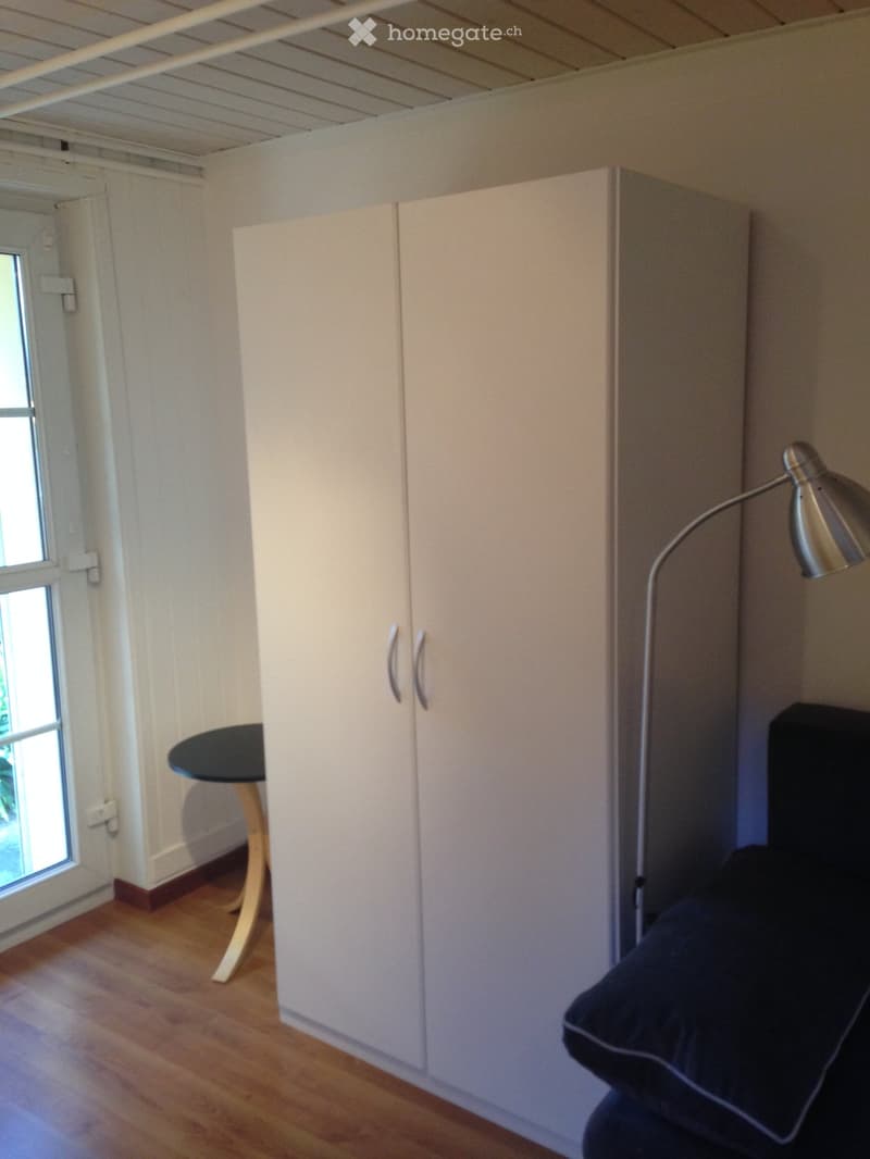 Room with white wardrobe, wooden floor, white walls, black round table, black chair with pillow, floor lamp.