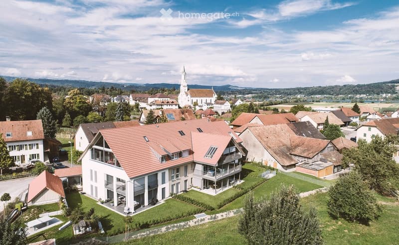 A modern two-story house with a red-tiled roof, large windows, and a spacious garden, situated in a rural village with other houses and a church in the background.