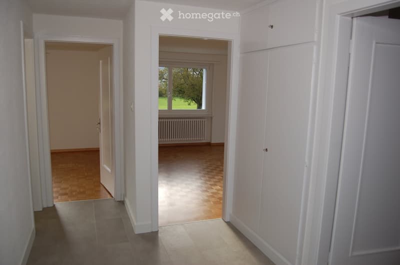 Empty hallway with white walls, tiled floor, and two open doors leading to adjacent rooms. One room has wooden flooring and a window with a view of greenery.
