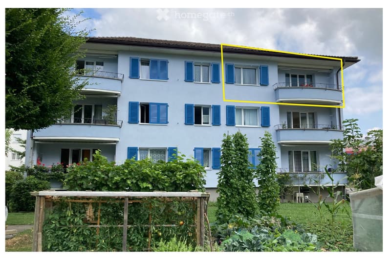 A three-story residential building with light blue walls, blue shutters, and multiple balconies. The building is surrounded by lush greenery and a garden area with vegetables and plants.