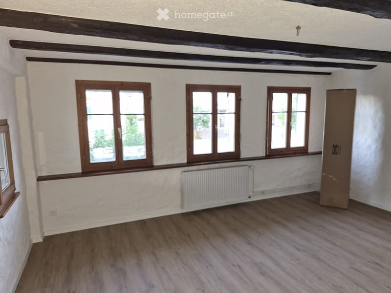 Empty room with wooden flooring, white walls, and three windows with wooden frames. A radiator is visible under the windows.
