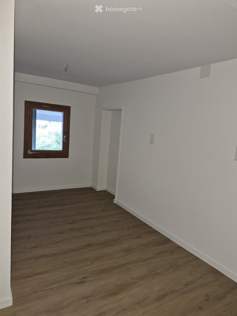 Empty room with wooden flooring, white walls, and a single window with a brown frame. There is a doorway leading to another space.