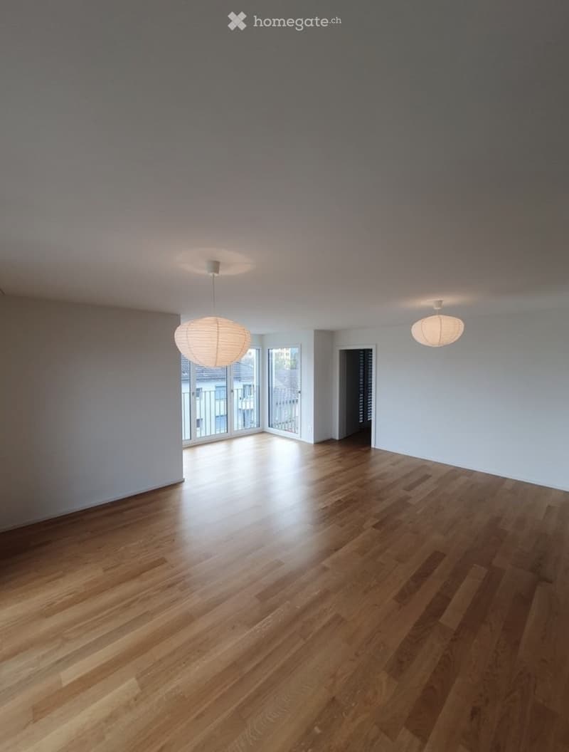 Empty room with wooden flooring, white walls, and two hanging lamps. There is a doorway leading to another room and a large window with a view of the outside.