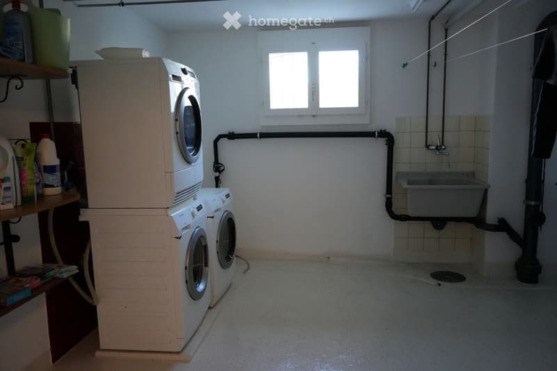 Two stacked washing machines, a sink, shelves with cleaning products, a window