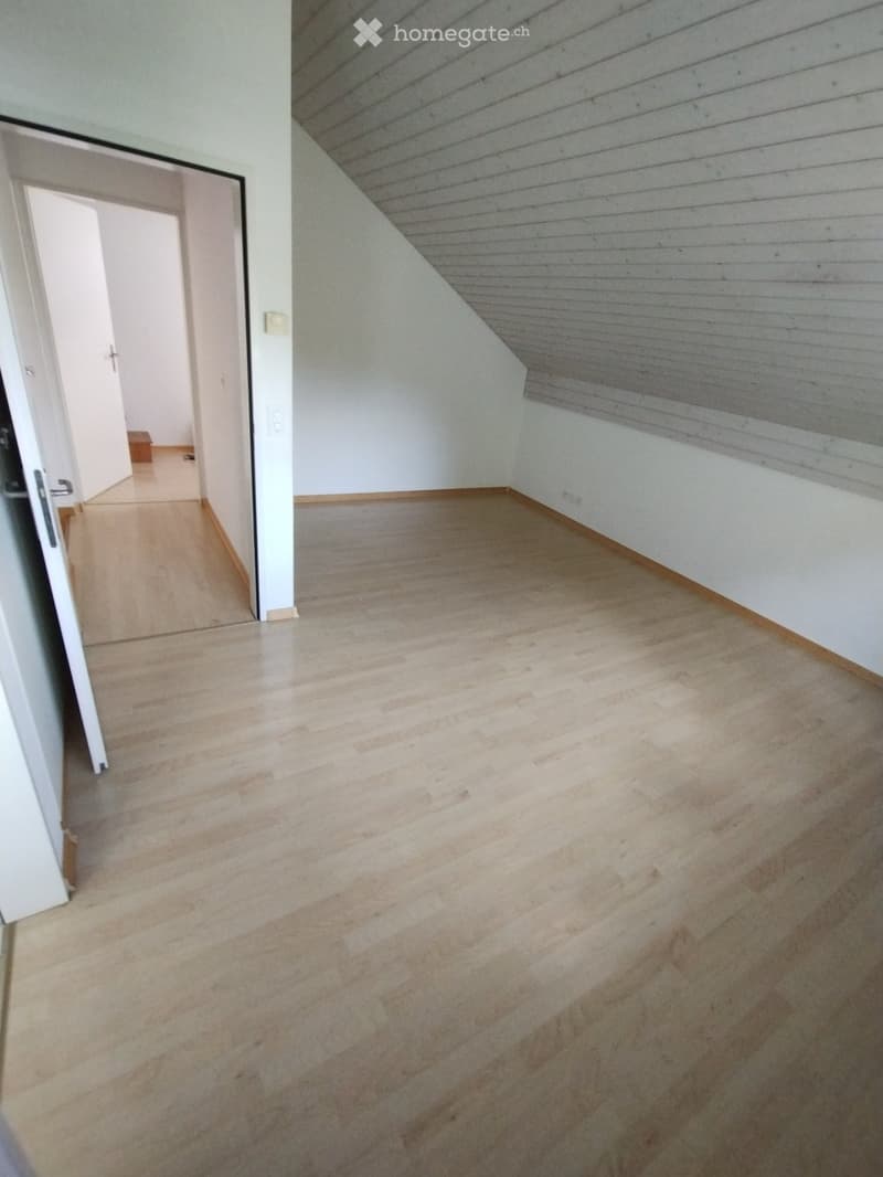 Empty room with wooden flooring, white walls, and a slanted ceiling. The room has a doorway leading to another space.
