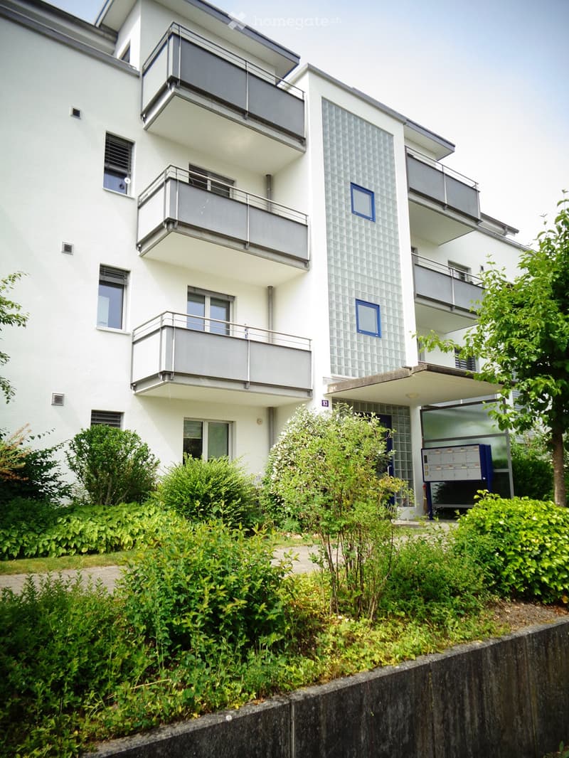 Modern white apartment building with multiple balconies, large windows, and surrounding greenery.