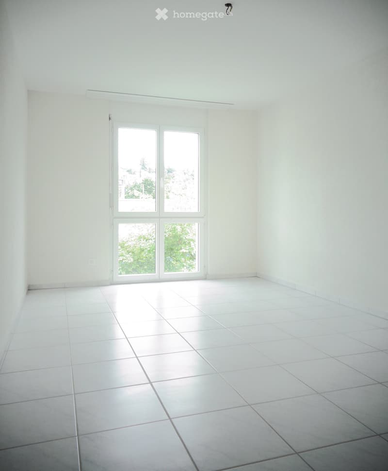 Empty room with white tiled flooring and white walls, featuring a large sliding glass door with a view of greenery outside.