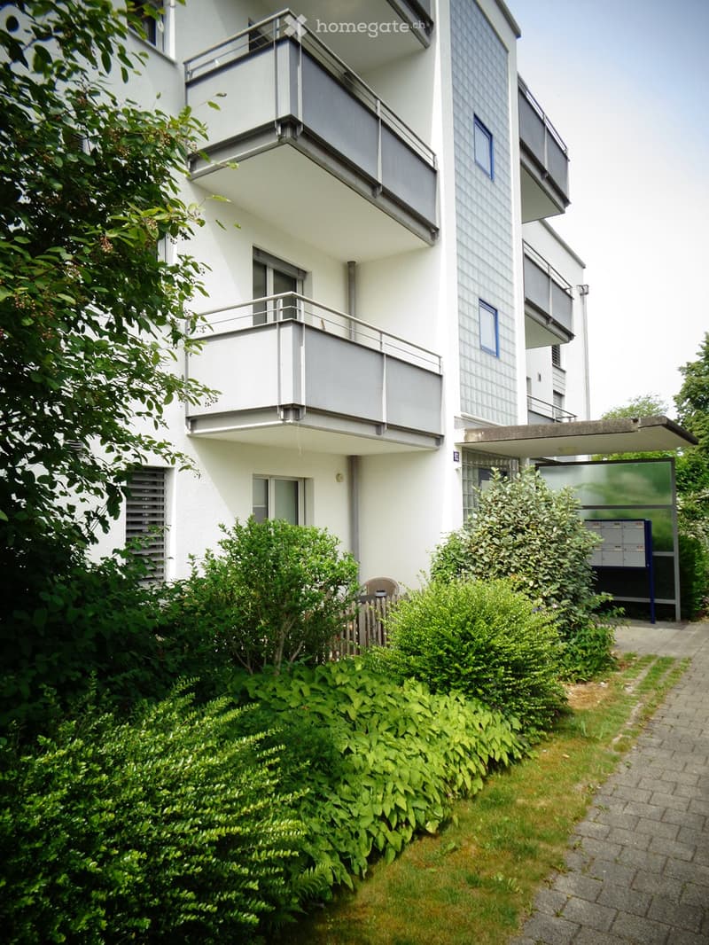 Modern multi-story apartment building with balconies, surrounded by greenery and a paved pathway.