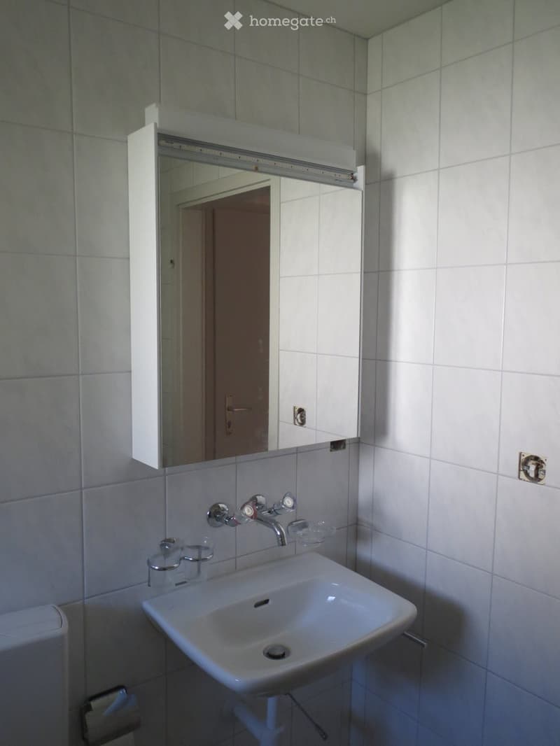 A bathroom with white tiled walls, a white toilet, a white sink with a faucet, a mirror, and a soap dish.