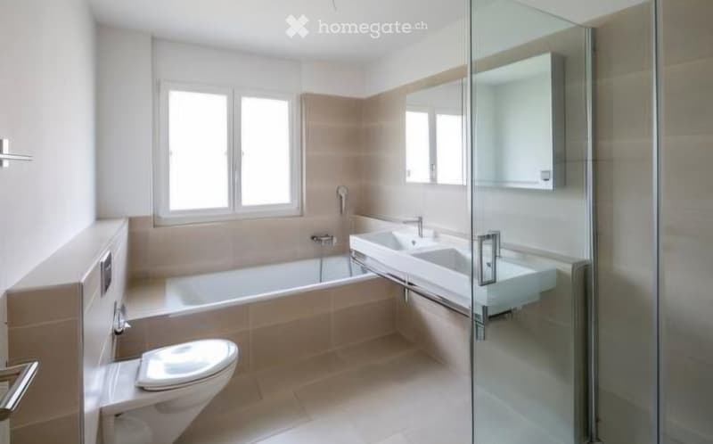 Modern bathroom with a bathtub, toilet, double sink, and a glass-enclosed shower area. Light-colored tiles and ample natural light from windows.