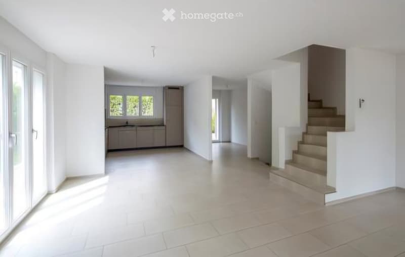 Empty hallway with white walls, tiled floor, and a staircase leading to an upper level. The hallway has a view into a kitchen area with cabinets and a sink.