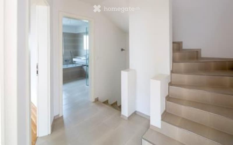 Bright hallway with white walls, wooden floor, and a staircase leading upwards. An open doorway reveals a bathroom with a bathtub and sink.