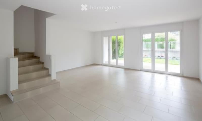 Empty room with white walls, tiled floor, and large glass doors leading to an outdoor area. A staircase is visible on the left side.