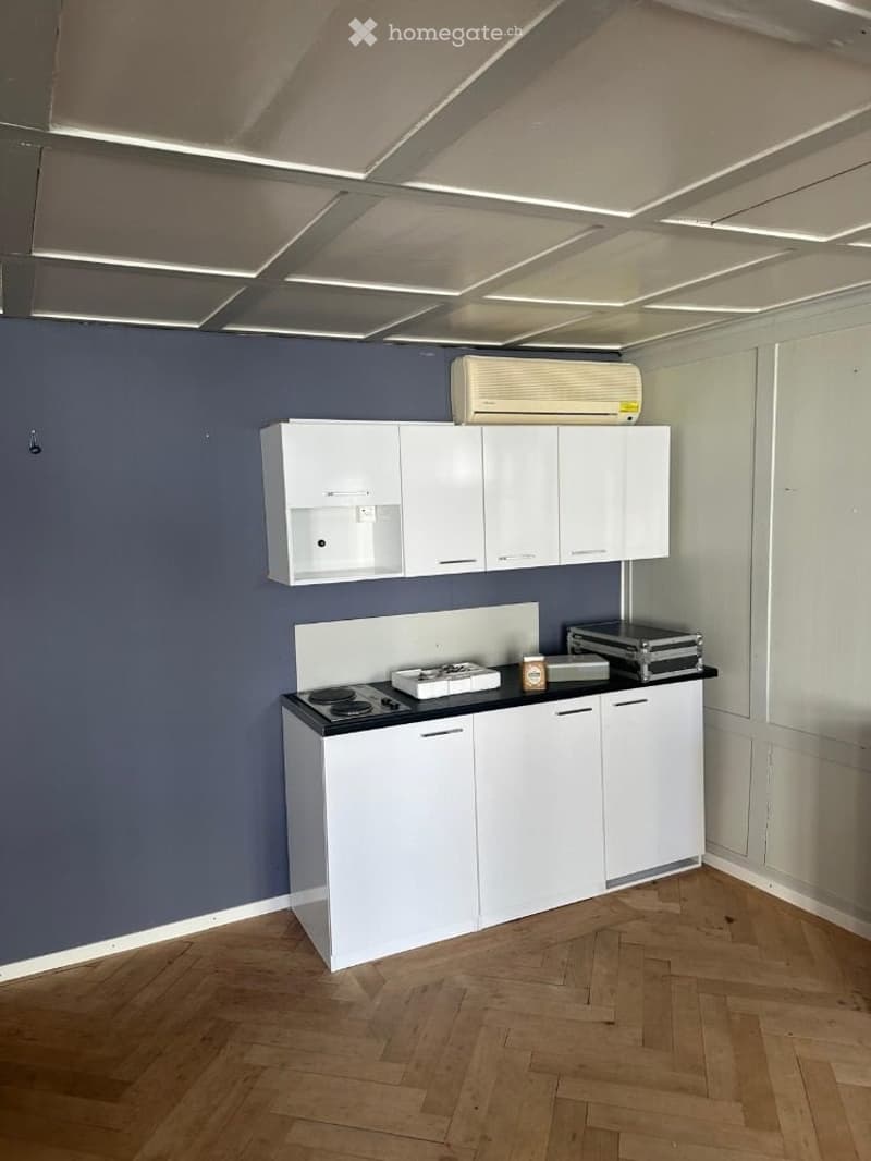 Modern kitchen with white cabinets, induction stove, and air conditioner above. Wooden floor and grey walls.