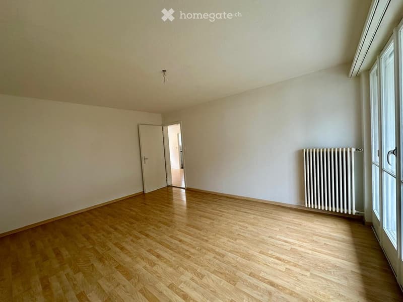 Empty room with wooden flooring, white walls, a radiator, and a door leading to another room.