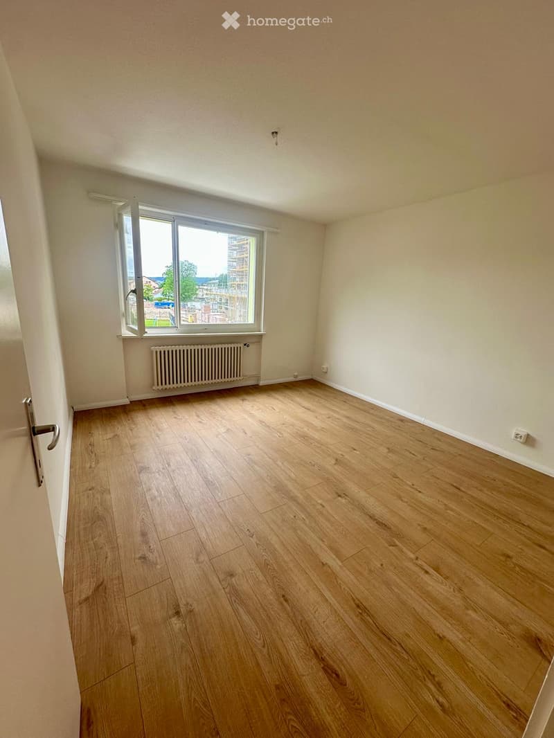 Empty room with wooden flooring, white walls, a window with a radiator below, and a door on the left side.