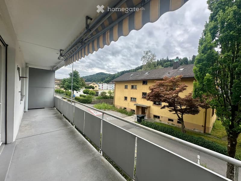 Balcony with metal railing, striped awning, overlooking a residential area with trees and houses.