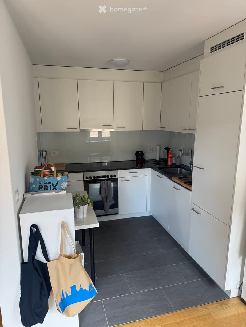 Modern kitchen with white cabinets, dark countertops, and stainless steel appliances. Includes an oven, sink, coffee maker, and a small table with a plant and handbags.
