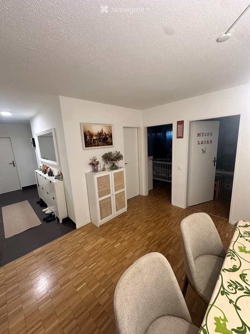 A well-lit hallway with wooden flooring, a white cabinet, a mirror, a picture frame, and a small table with a vase and decorative items. A sofa and chair are visible in the foreground.