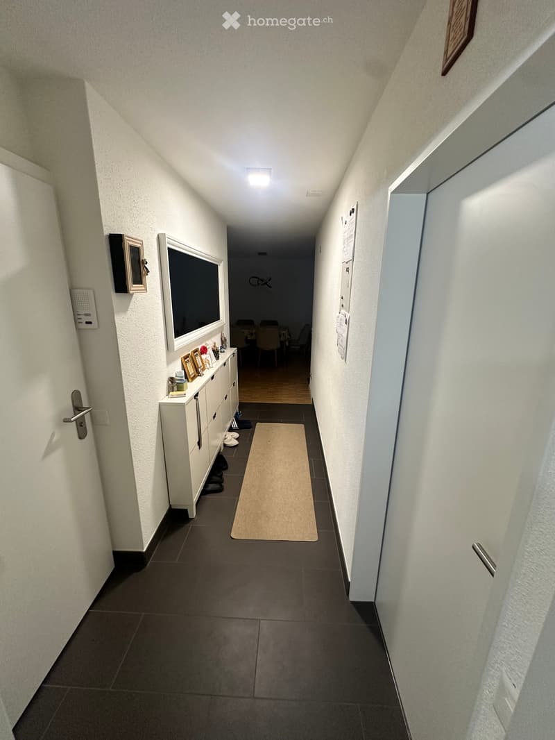 A narrow hallway with white walls, dark tiled floor, and a small rug. A white cabinet with drawers and framed pictures is on the left, and a partially visible room with chairs and a table is at the end.
