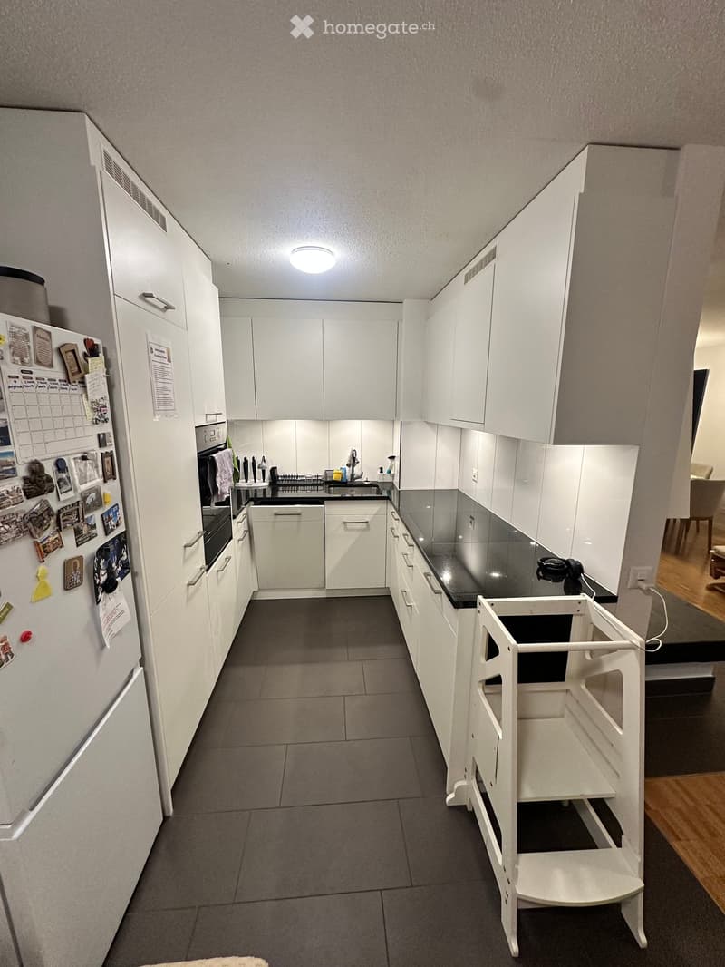 Modern kitchen with white cabinets, black countertop, stainless steel appliances, and a small white stool near the counter.