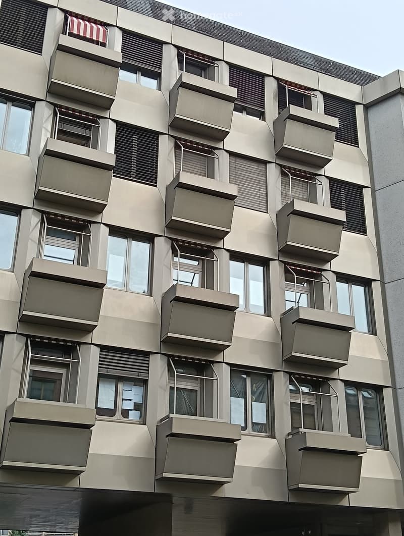 Modern multi-story building with numerous balconies and large windows, featuring a clean, geometric design with beige and grey tones.