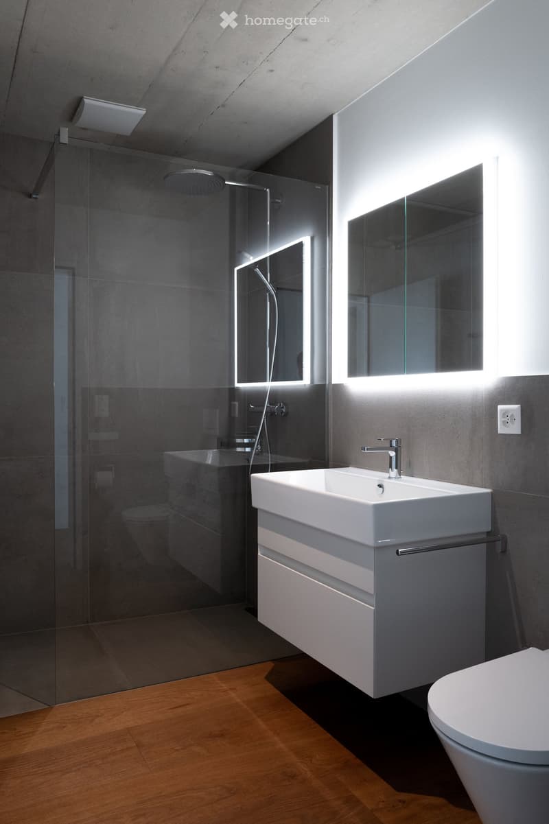 Modern bathroom with a white sink, chrome faucet, large mirror with built-in lighting, glass-enclosed shower, and a toilet. The floor is wooden, and the walls are tiled in grey.