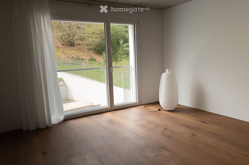 Empty room with wooden flooring, large sliding glass doors leading to a balcony, white walls, and a single white decorative lamp.
