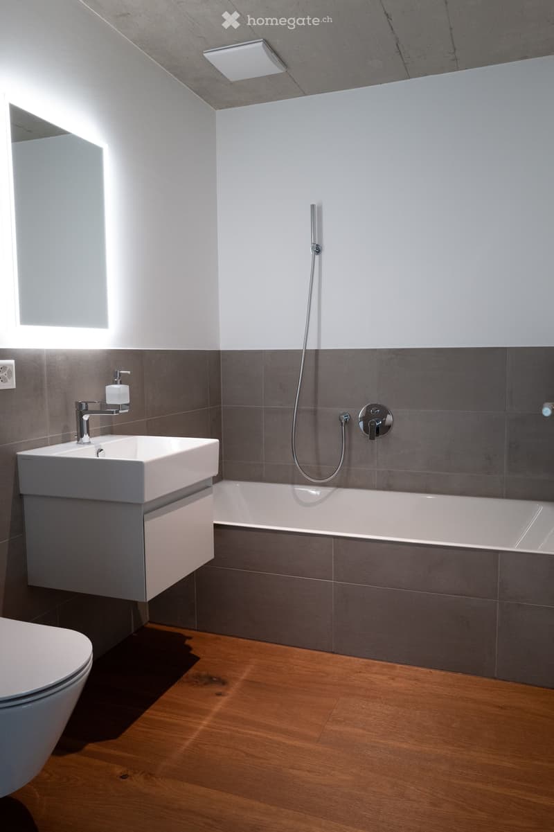 Modern bathroom with a white sink, bathtub, and toilet. Grey tiles on lower walls, wooden flooring, and sleek fixtures.