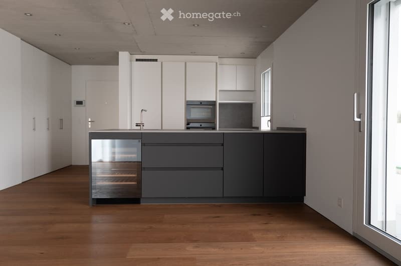 Modern kitchen with white cabinets, dark grey lower cabinets, a central island, and a wine cooler. Features a sleek, minimalist design with a wooden floor and large windows.