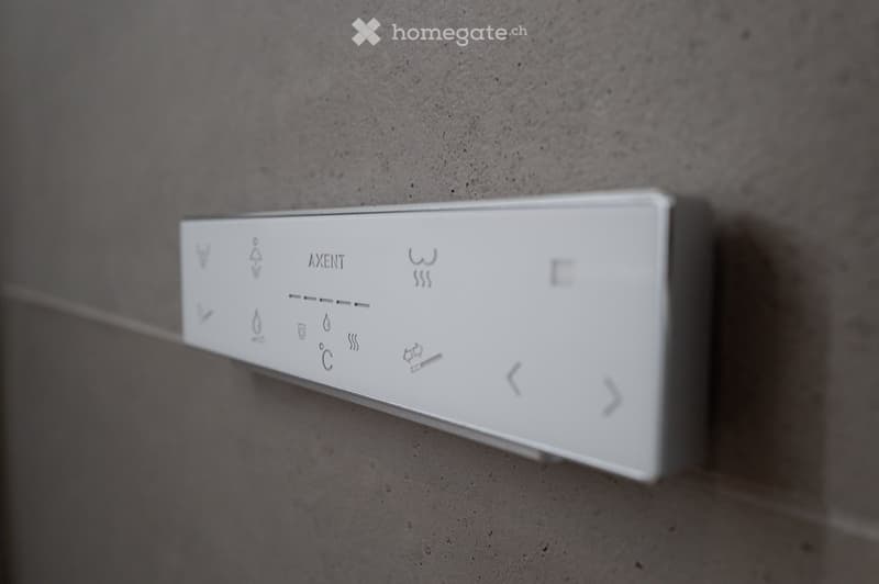 A modern, sleek white control panel with various icons and symbols mounted on a grey wall, likely for a bathroom or shower system.