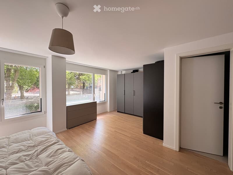 Modern bedroom with light wooden flooring, white walls, large windows, a bed with white bedding, a wardrobe, a cabinet, and a pendant light.