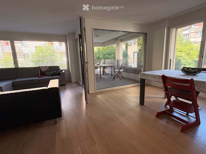 Living room with hardwood floors, large windows, and a balcony. Features a grey sectional sofa, a dining table with chairs, and a bowl on the table.