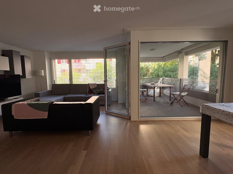 Bright living room with wooden flooring, large windows, and sliding glass doors leading to a balcony. Features a grey sectional sofa, a chair, a small table, and a television.