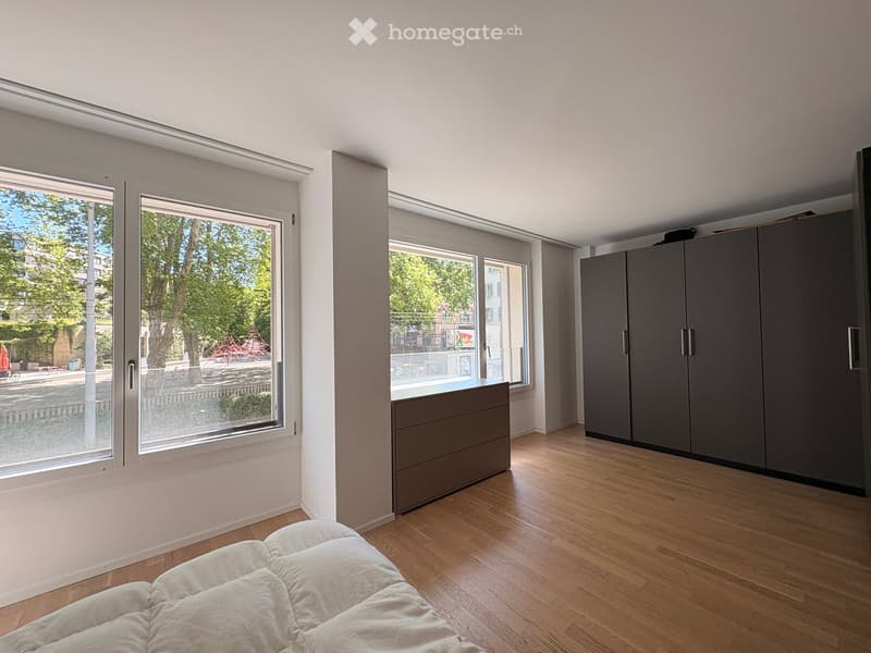 Bright bedroom with wooden flooring, large windows, a bed with white bedding, a wardrobe, and a drawer unit.