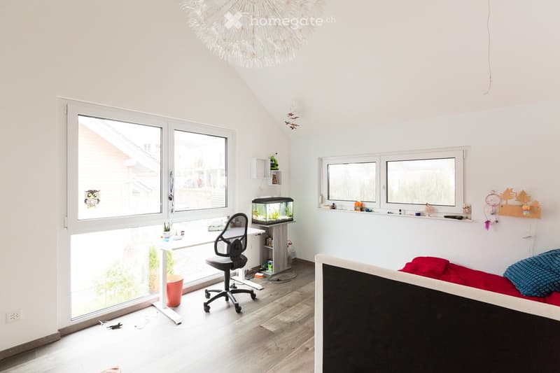 Bright, modern room with large windows, a desk area with a chair and fish tank, and a bed with pillows and a blanket.