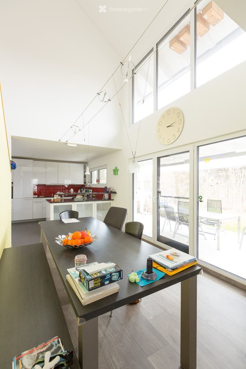 A modern, open-plan kitchen and dining area with a large dining table, chairs, a clock on the wall, and a kitchen area with white cabinets and a red backsplash.