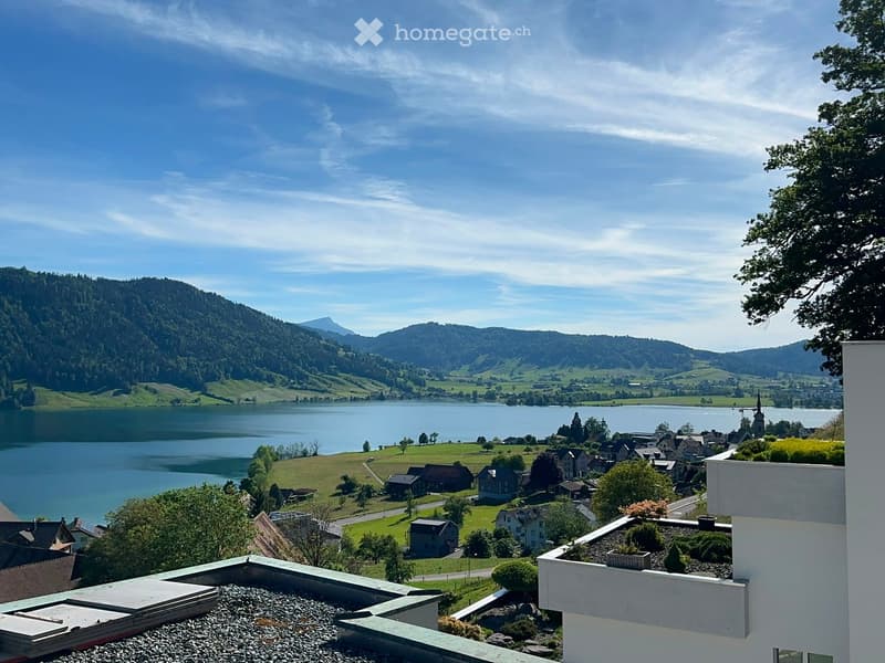 Panoramic view from a balcony overlooking a serene lake surrounded by lush green hills and a small village with scattered houses and trees.
