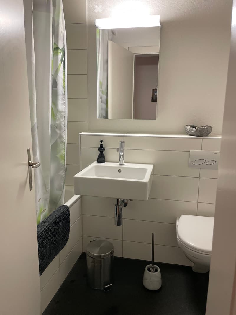 Modern bathroom with white tiles, a white sink, a chrome faucet, a toilet, a mirror with built-in lighting, a towel, a soap dispenser, and a trash can.