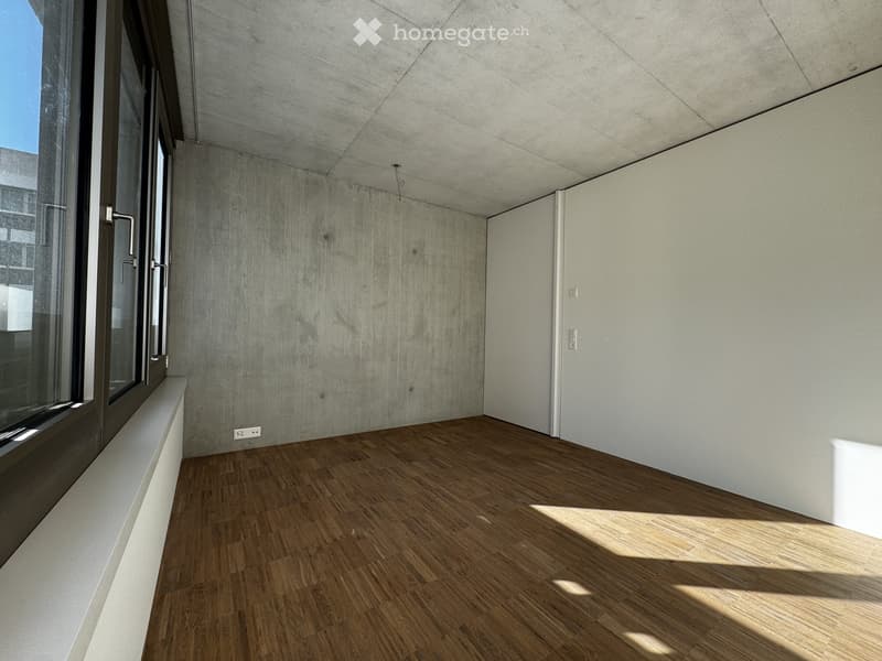 Empty room with wooden flooring, large window, and concrete walls.