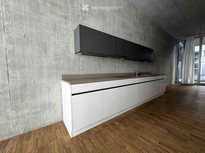 Modern kitchen with white lower cabinets, grey upper cabinets, a concrete countertop, and a sink with a faucet. The walls are textured concrete, and the floor is wooden.