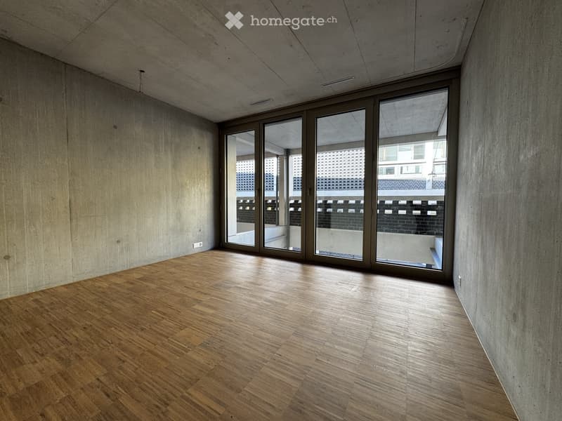 Empty room with wooden flooring and large glass sliding doors leading to a balcony. Bare concrete walls and ceiling.