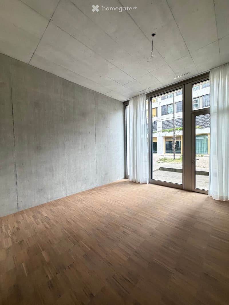 Empty room with wooden flooring, concrete walls, large windows with white curtains, and a sliding glass door leading to an outdoor area.