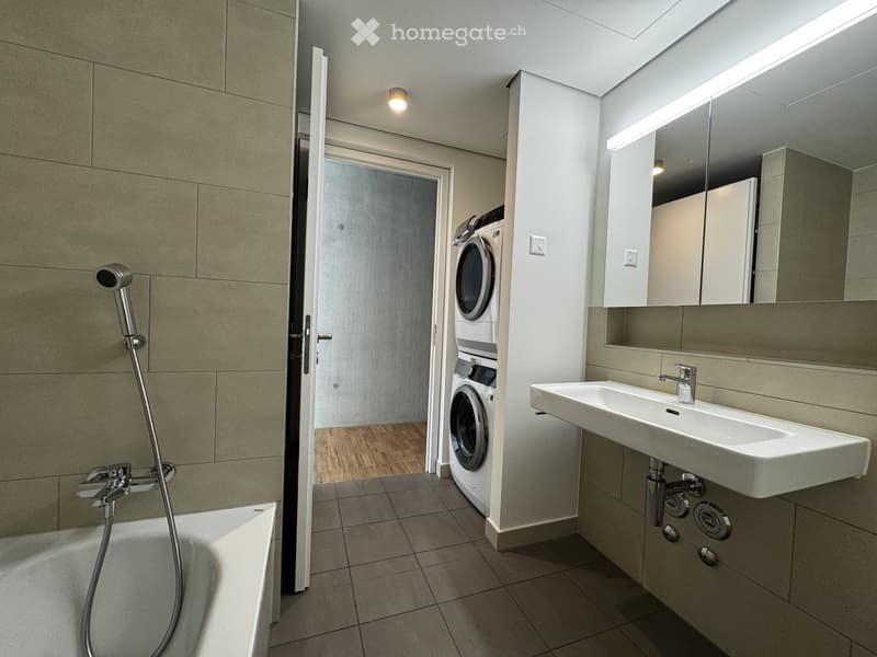 Modern bathroom with a bathtub, sink, mirror, and a washing machine in an adjacent space.