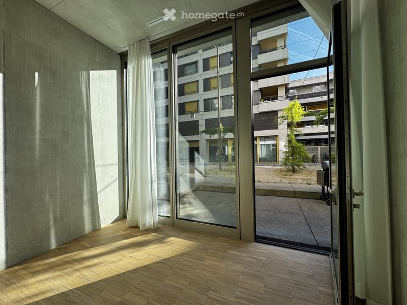Empty room with wooden flooring, large glass sliding doors, and white curtains. Sunlight streams in, casting shadows on the floor.