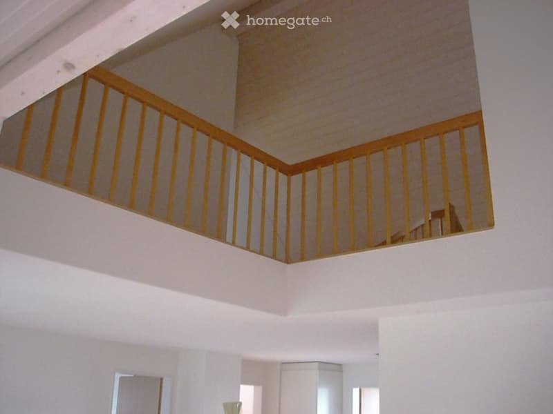 A modern hallway with white walls and a wooden staircase railing, featuring a raised ground floor and ample natural light from windows.