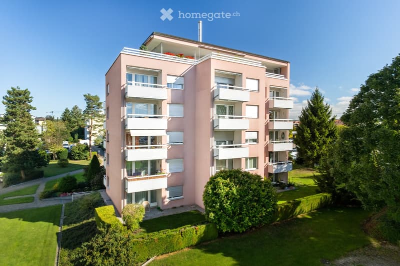 A multi-story apartment building with a pink exterior and white balconies. The building is surrounded by lush greenery and landscaping, including trees and shrubs.